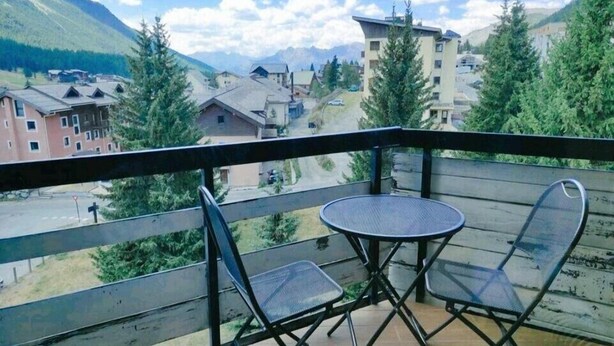 Un balcon avec deux chaises et une table, d’où l’on peut admirer un village avec ses bâtiments et, au loin, des montagnes.