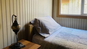 A bedroom with a bed, a nightstand, a lamp, and a window with blinds. - Hostel Gran Via (Punta Arenas)