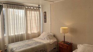 A single bed with a patterned bedspread, a bedside table with a lamp, and a window with sheer curtains. - Hostel Gran Via (Punta Arenas)