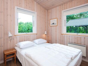 Un dormitorio con una cama, dos lámparas de noche, una mesita de noche de madera, un cuadro enmarcado y una ventana con vistas a zonas verdes.