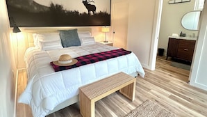 A bedroom with a large bed, a wooden bench, a rug, a wall-mounted picture of a deer, and a nightstand with a lamp. - Adventure Basecamp Fly Fishing Modern Alpine Home Near Jackson “Superfly” Cabin (Alpine)