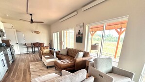 A spacious living area with a large sectional sofa, a coffee table, and a dining area with chairs. There is a kitchen area with cabinets and a refrigerator. The room has large windows and a ceiling fan. - Adventure Basecamp Fly Fishing Modern Alpine Home Near Jackson “Superfly” Cabin (Alpine)