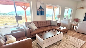 A spacious living room with a brown leather sofa, a white ottoman, and a wooden coffee table. Large windows offer a view of a field and mountains. - Adventure Basecamp Fly Fishing Modern Alpine Home Near Jackson “Superfly” Cabin (Alpine)