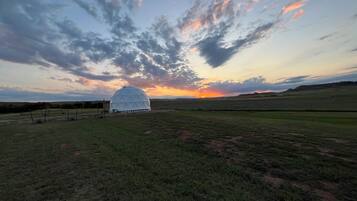 Une structure en forme de dôme géodésique se dresse dans un champ verdoyant, avec un coucher de soleil en arrière-plan.