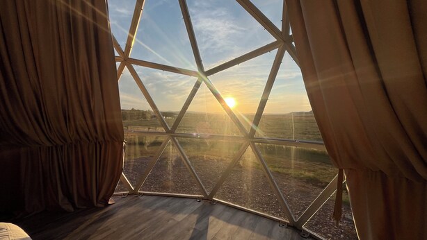 A room with a large window featuring a geometric metal frame, a wooden floor, and curtains. - WYnDome - Glamping Dome in Beulah, WY (Beulah)