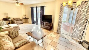 A living room with a sofa, coffee table, TV, and a balcony with patterned curtains. - Spacious House w/ Heated Pool & Hot Tub near Tampa (Brandon)