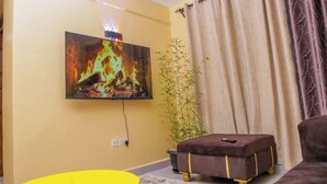A living room with a flat-screen TV displaying a fire, a yellow table, a brown ottoman, and a bamboo plant. - Lux Suites The Urbanite Apartments (Kitengela)