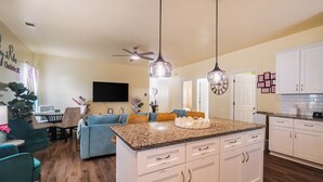 A modern kitchen with white cabinets, granite countertops, and a central island. There is a dining area with a blue sofa and a TV mounted on the wall. - 3BR Getaway Hot Tub, Games & Family Fun (Charlotte)