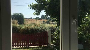 Udsigt gennem et glasdør til et landligt landskab med rødt hegn og grønt vegetation.