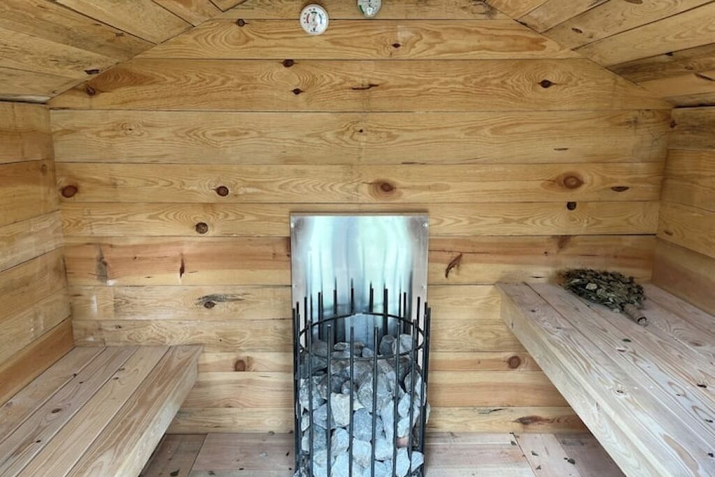 L’intérieur d’un chalet en bois, avec une cheminée en métal entourée de bûches, des bancs en bois et un plafond en bois avec des poutres apparentes.