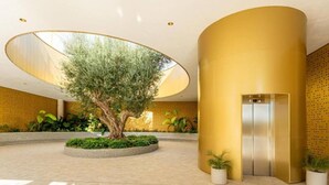 Un hall d’entrée spacieux avec un grand arbre, un ascenseur central et un mur jaune orné d’un motif décoratif.