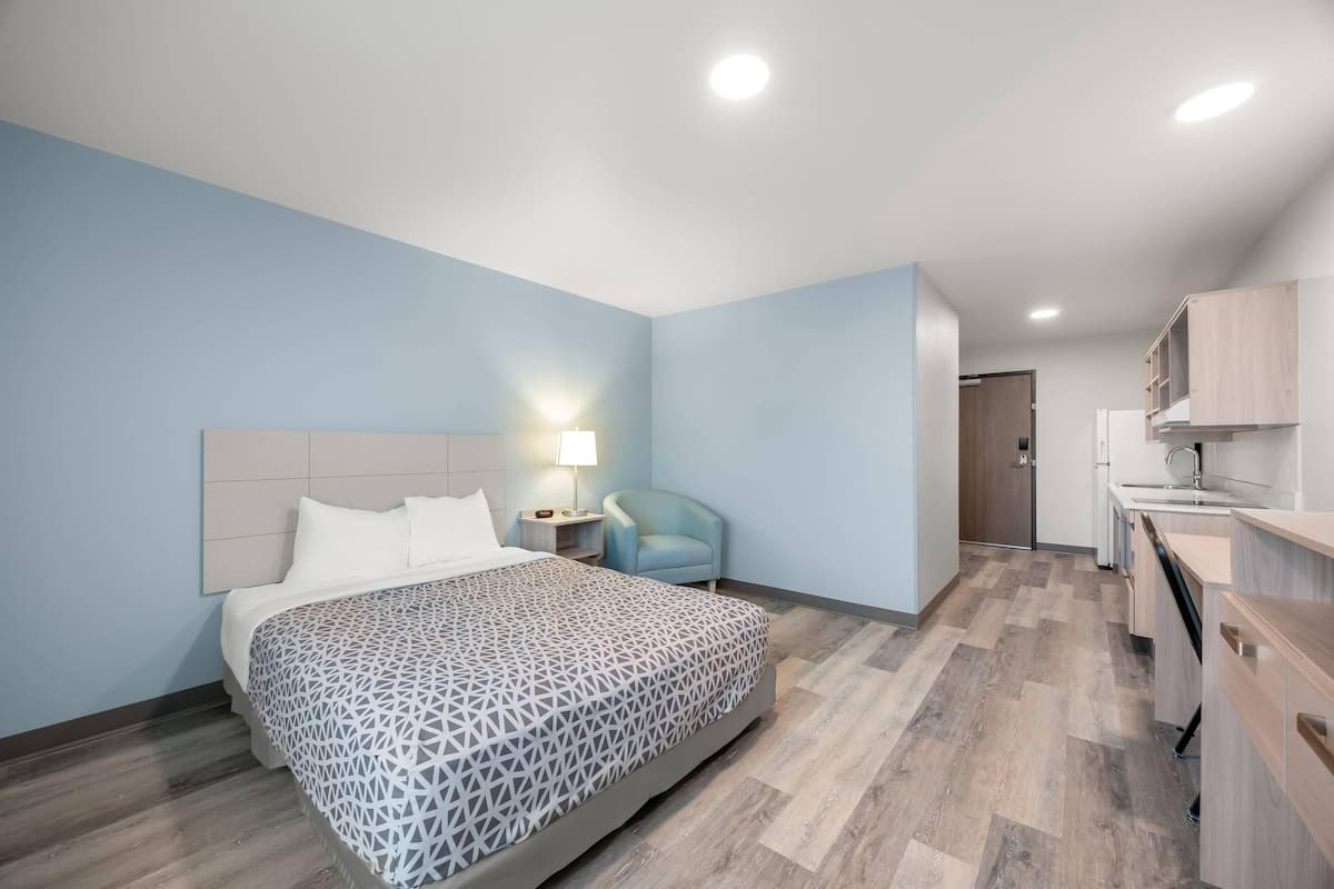 Een moderne hotelkamer met een groot bed, een nachtkastje met lamp, een stoel en een kitchenette.