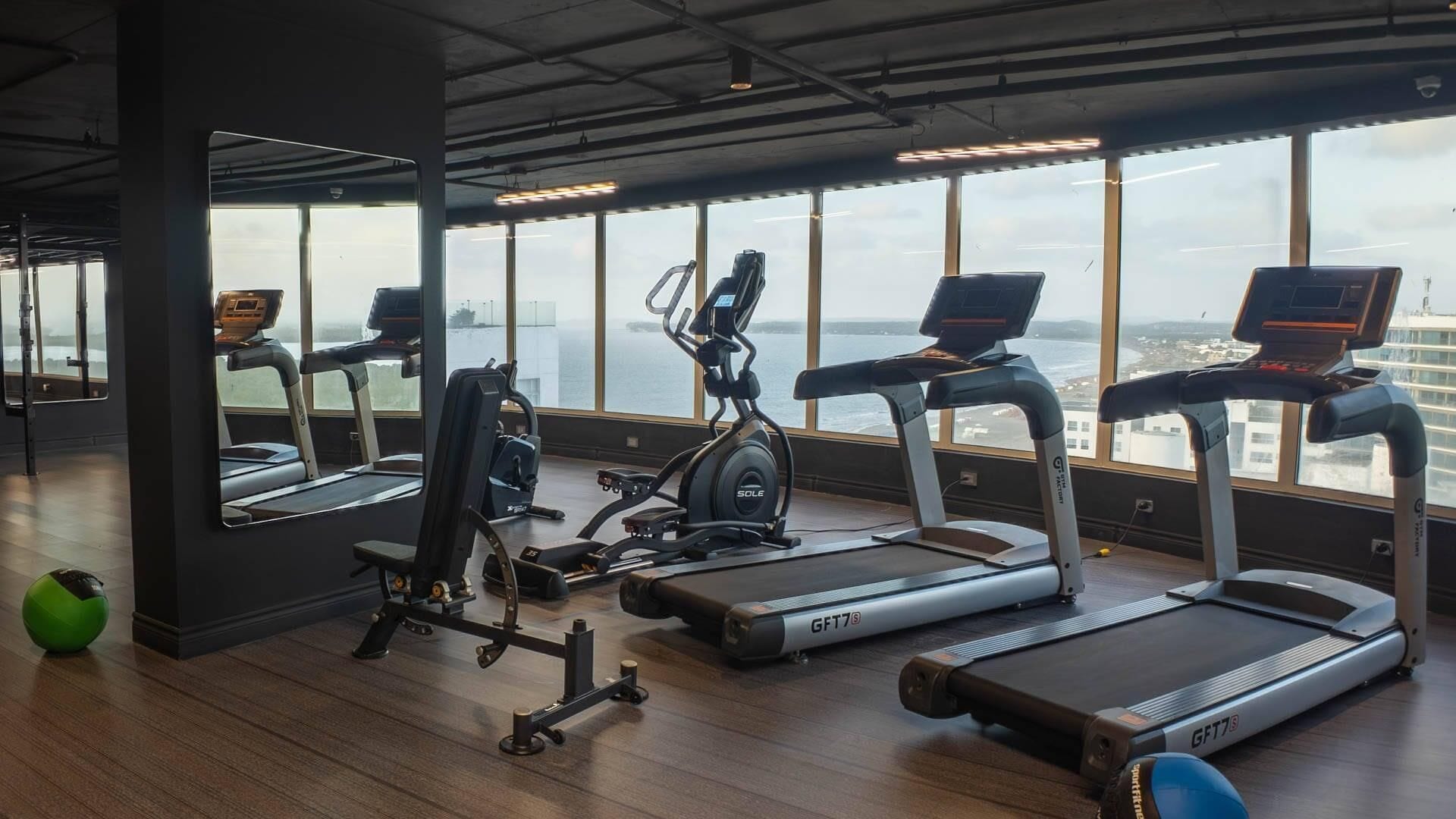 A gym with treadmills, an elliptical, and a rowing machine, with large windows overlooking a cityscape.