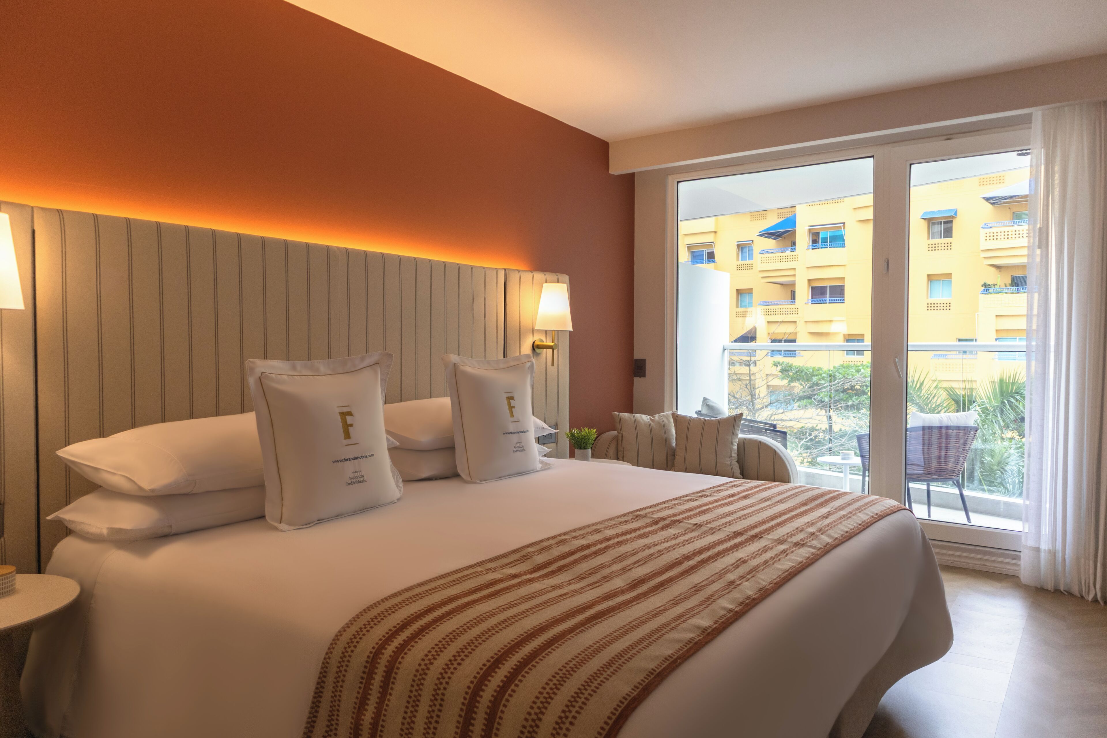 A hotel room with a large bed, two bedside lamps, a view of a building through a sliding door, and a small table with a chair.