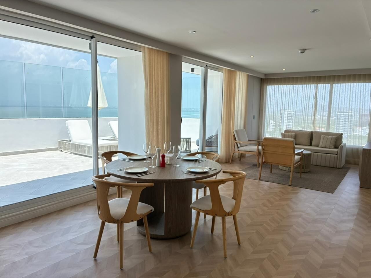 Uma sala de estar moderna com uma mesa de jantar redonda, cadeiras claras, um sofá e janelas amplas com vista para o mar.