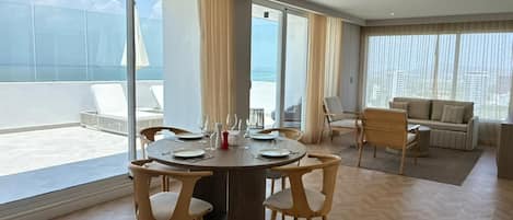 Una sala moderna con una mesa redonda de comedor, sillas de colores claros, un sofá y ventanas amplias con vistas al mar.