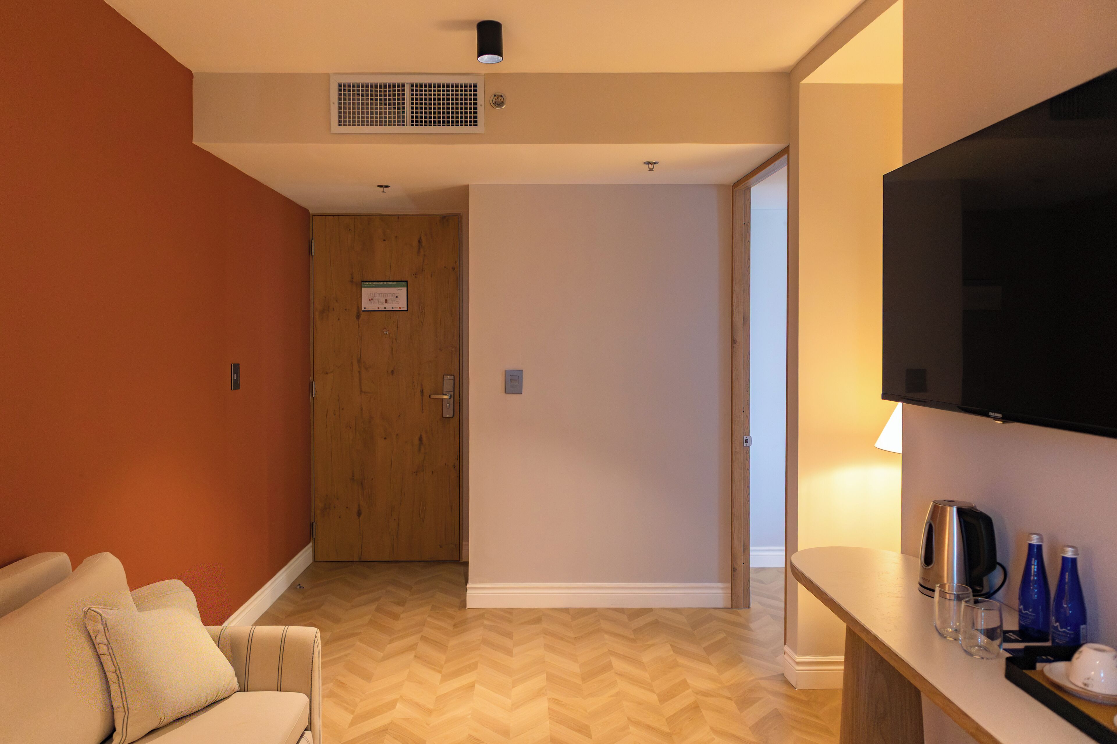 A hotel room with a wooden door, a flat-screen TV, a sofa, and a coffee table with glasses and a kettle.
