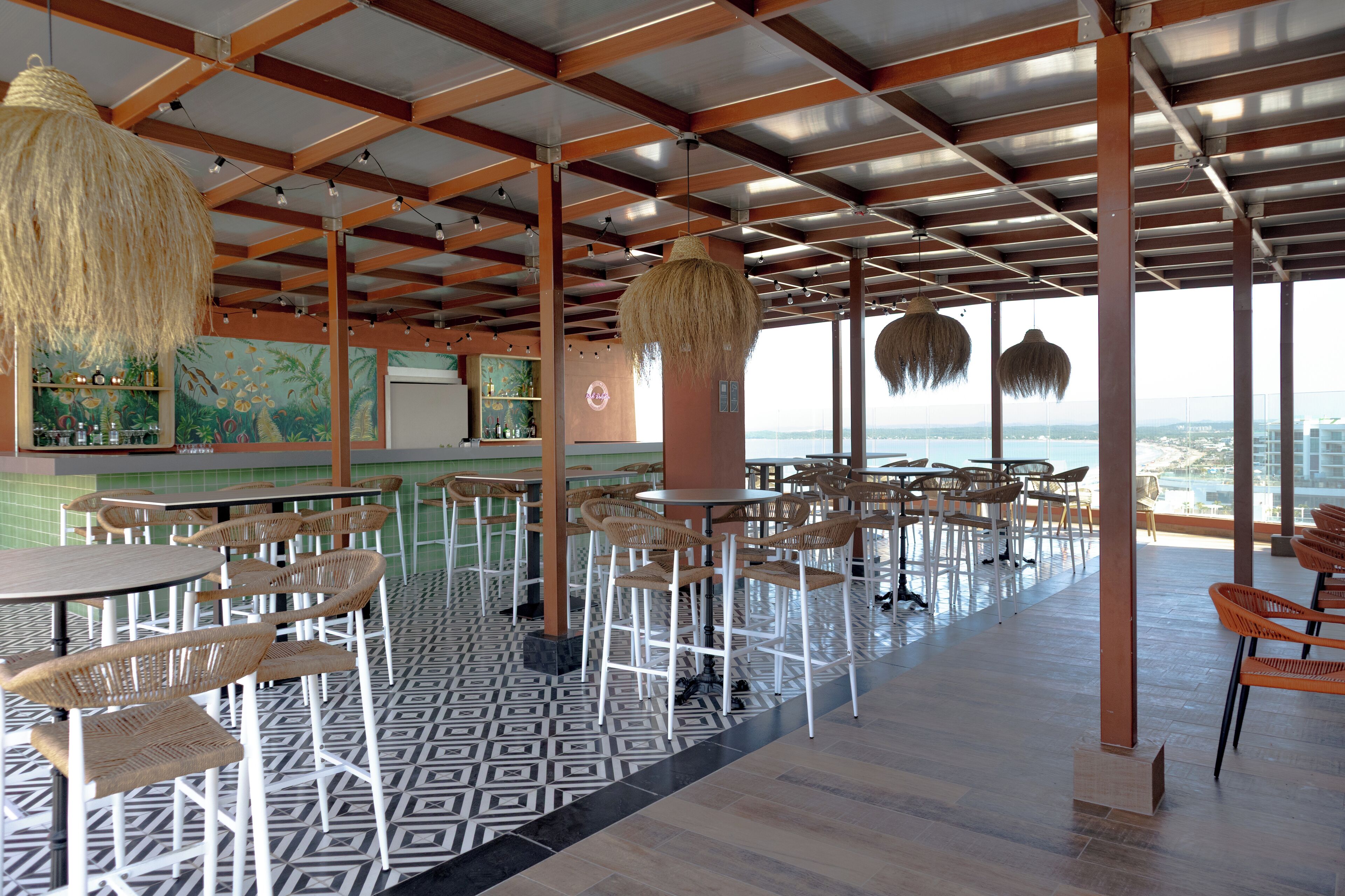 A spacious indoor area with high ceilings, wooden beams, and a patterned floor. Tables and chairs are arranged for dining, and a large window offers a view of the city skyline.