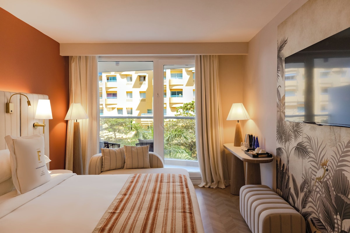 A hotel room with a large bed, two bedside lamps, a small table with a tray, a bench, and a large window with curtains.