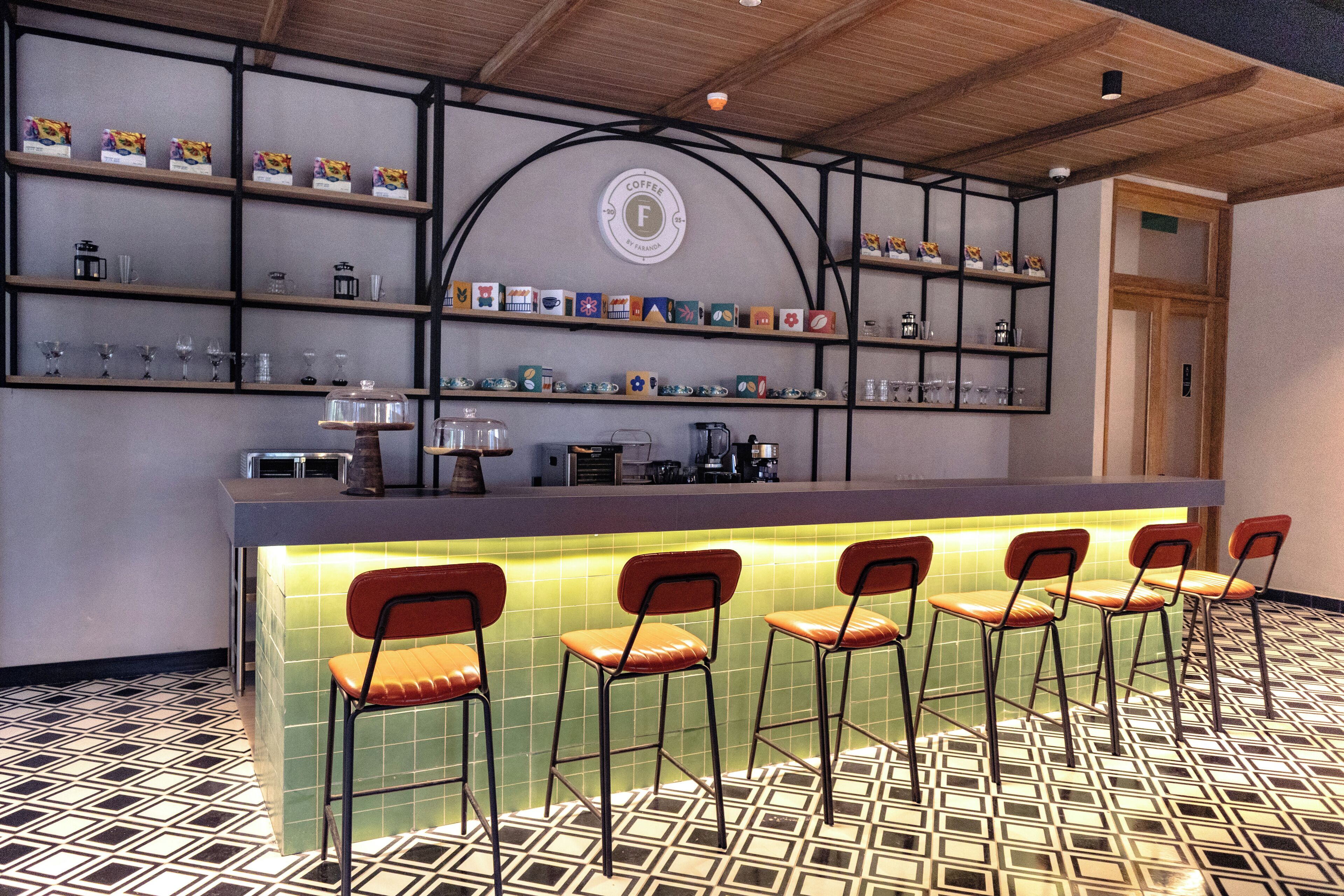 Una zona de bar con suelo de baldosas, una barra con taburetes y estantes con diversos objetos.