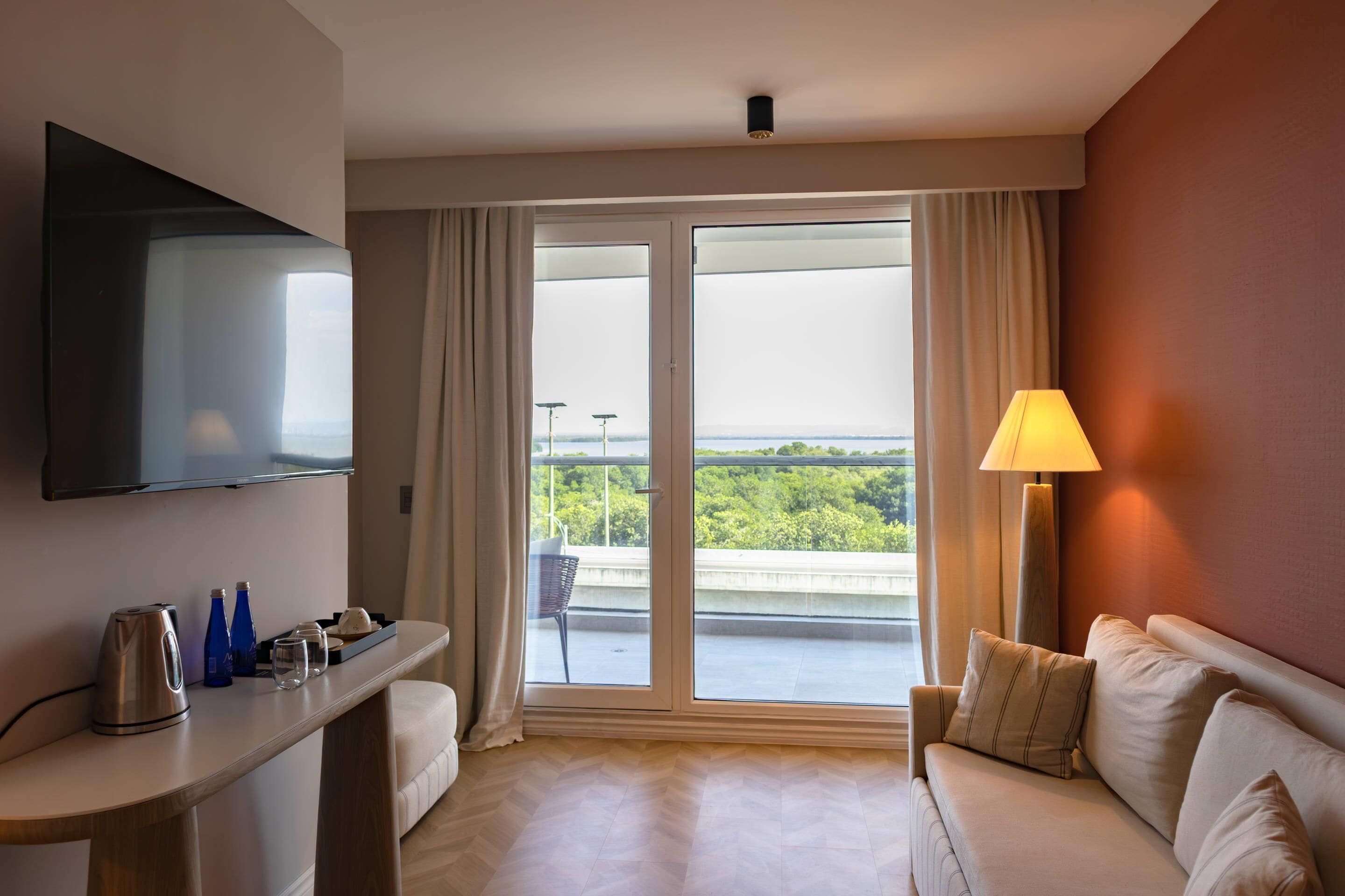 Habitación de hotel con sofá, televisor de pantalla plana, mesa con tetera y botellas, y balcón con vistas.