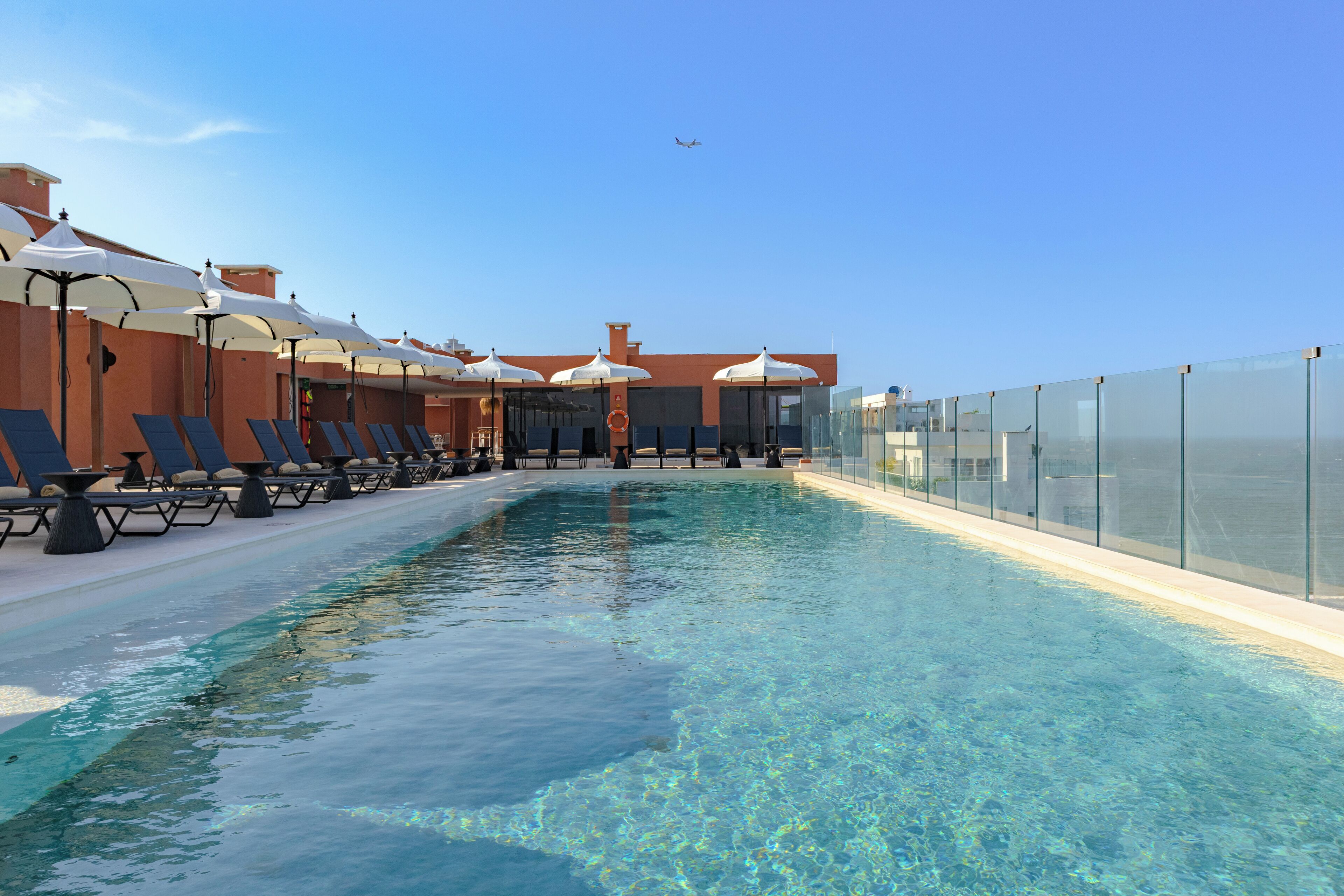 A swimming pool with lounge chairs and umbrellas, surrounded by a glass fence.