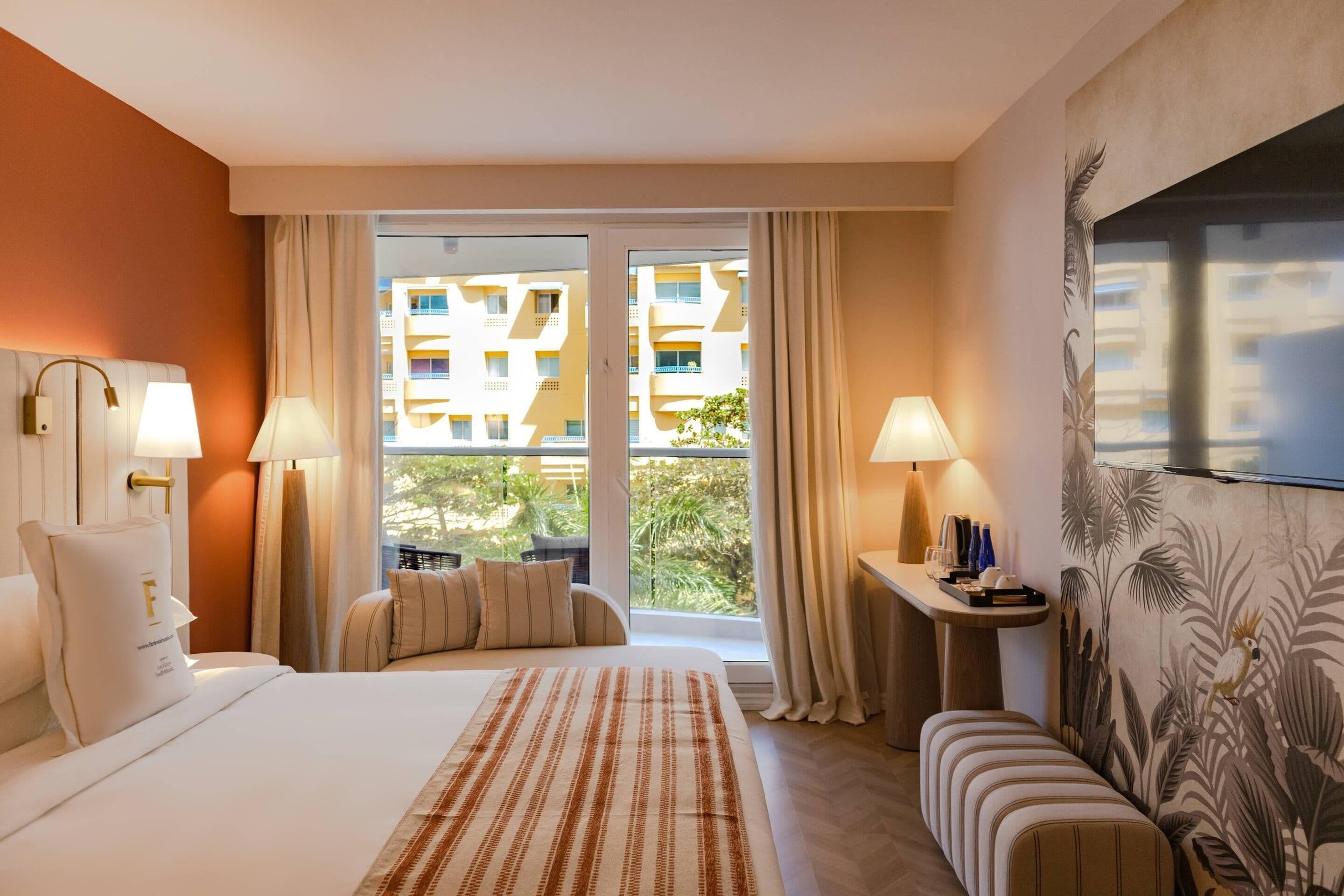 Une chambre d’hôtel avec un grand lit, deux lampes de chevet, une fenêtre avec des rideaux, une petite table avec un plateau, et un mur orné d’une fresque tropicale.
