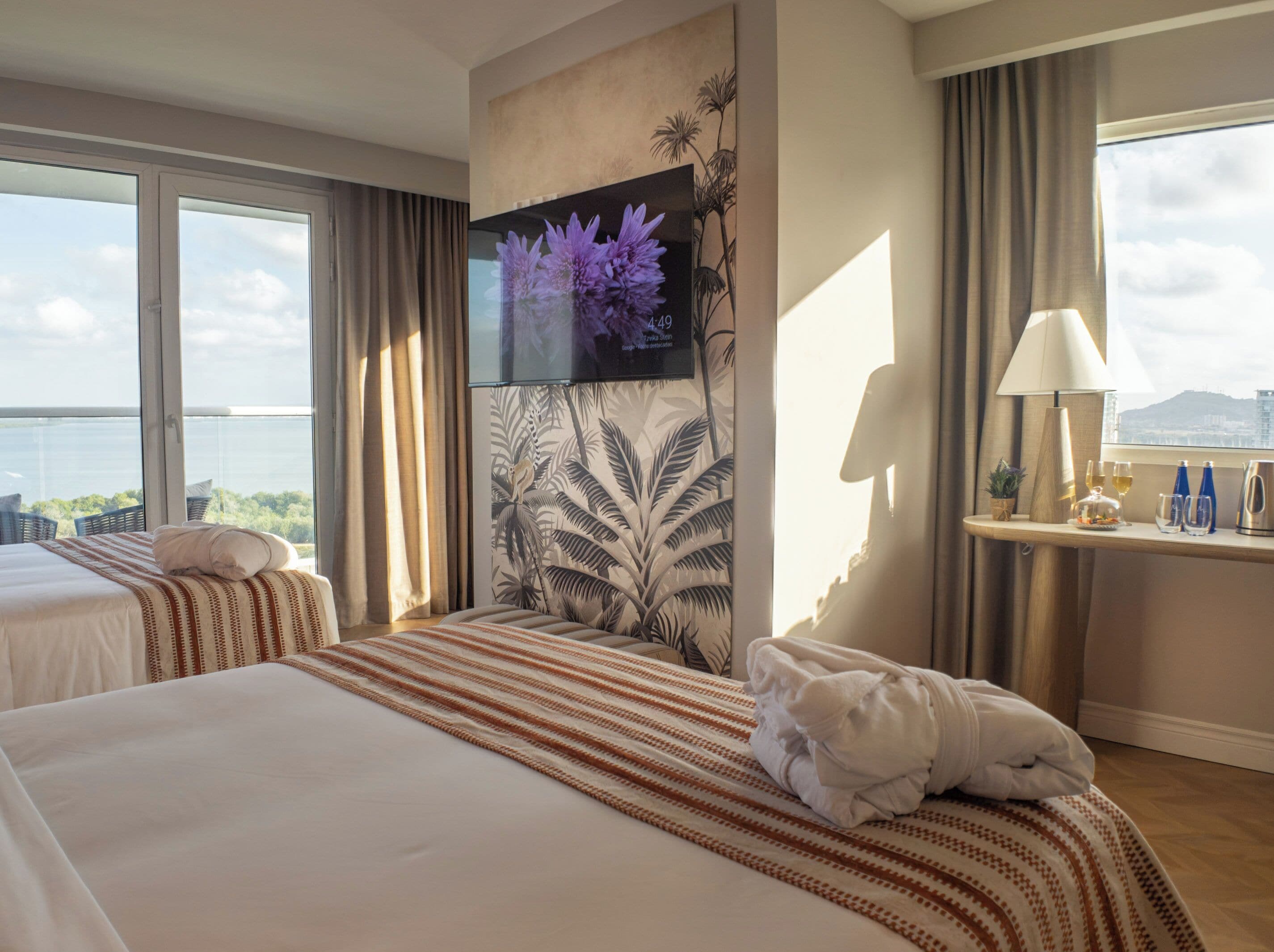 Habitación de hotel con dos camas, un televisor montado en la pared, una ventana con vistas a la ciudad y una mesita de noche con una lámpara y vasos.