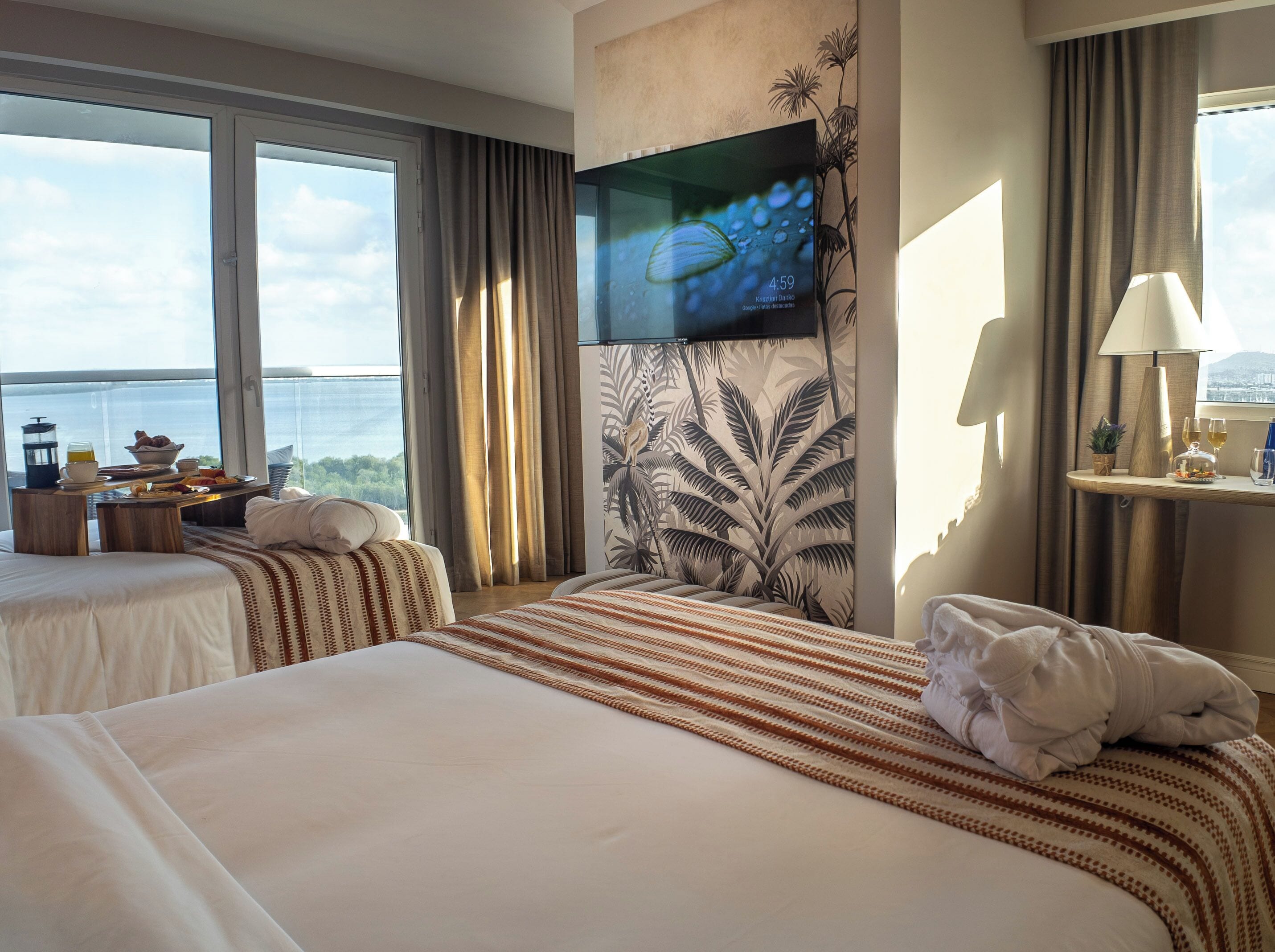 Une chambre d’hôtel avec deux lits, une télévision, une fenêtre donnant sur la mer, une petite table avec des articles pour le petit-déjeuner et une lampe.