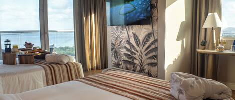 Habitación de hotel con dos camas, televisor, ventana con vista al mar, una mesita con artículos de desayuno y una lámpara.