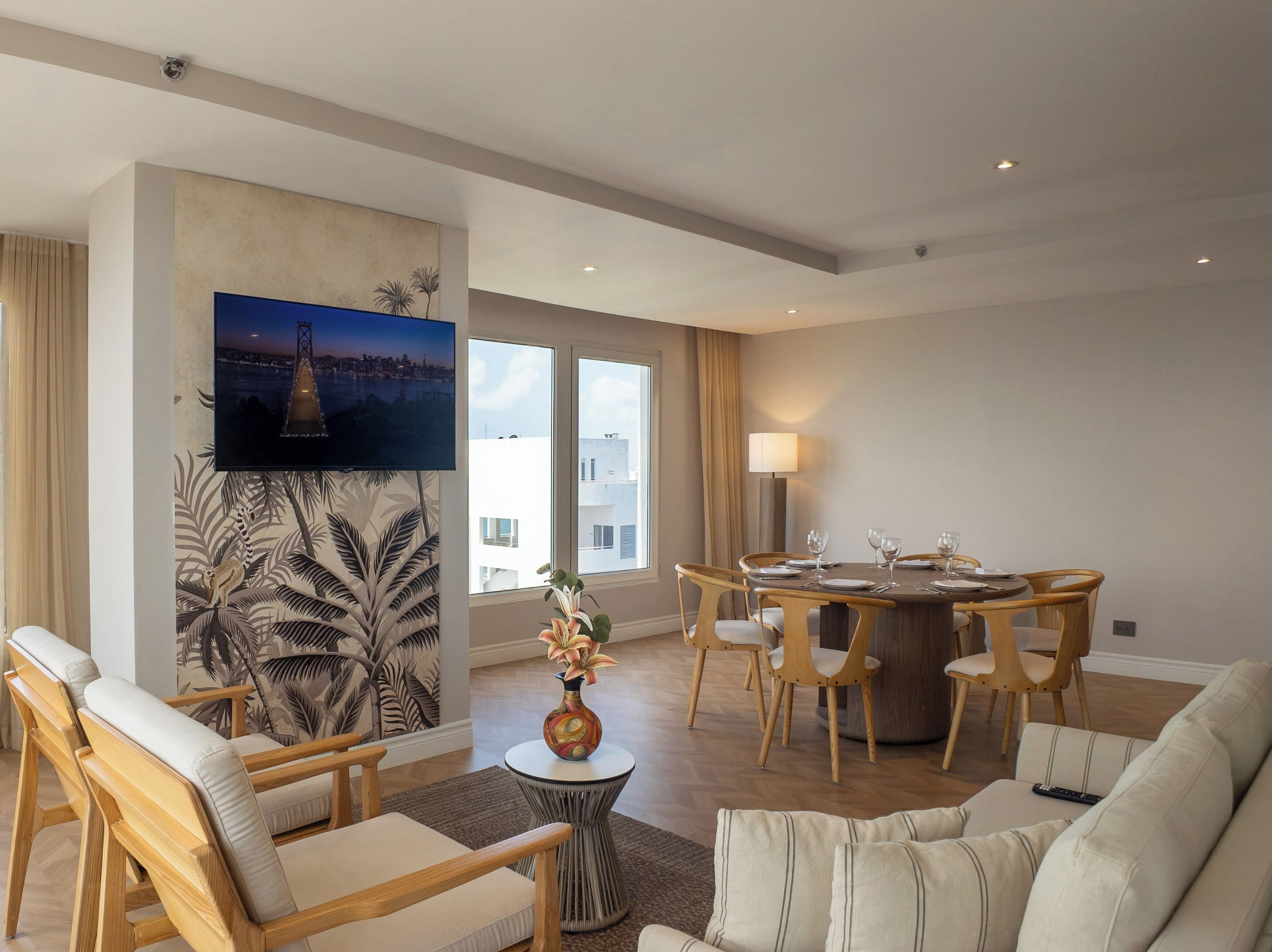 A modern living room with a flat-screen TV, a dining area with a round table and chairs, and a large window with a view of buildings.