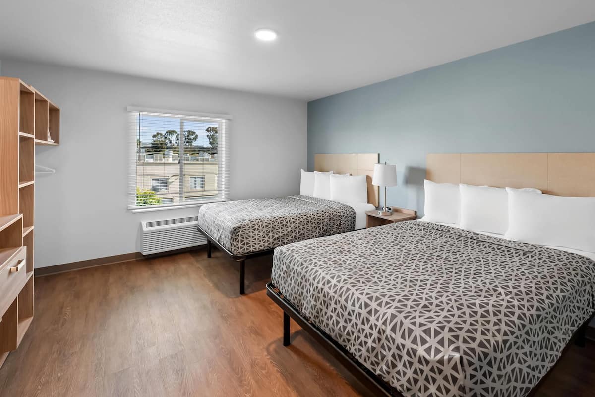 A hotel room with two beds, a window with blinds, a heater, a bedside table with a lamp, and a wooden wardrobe.