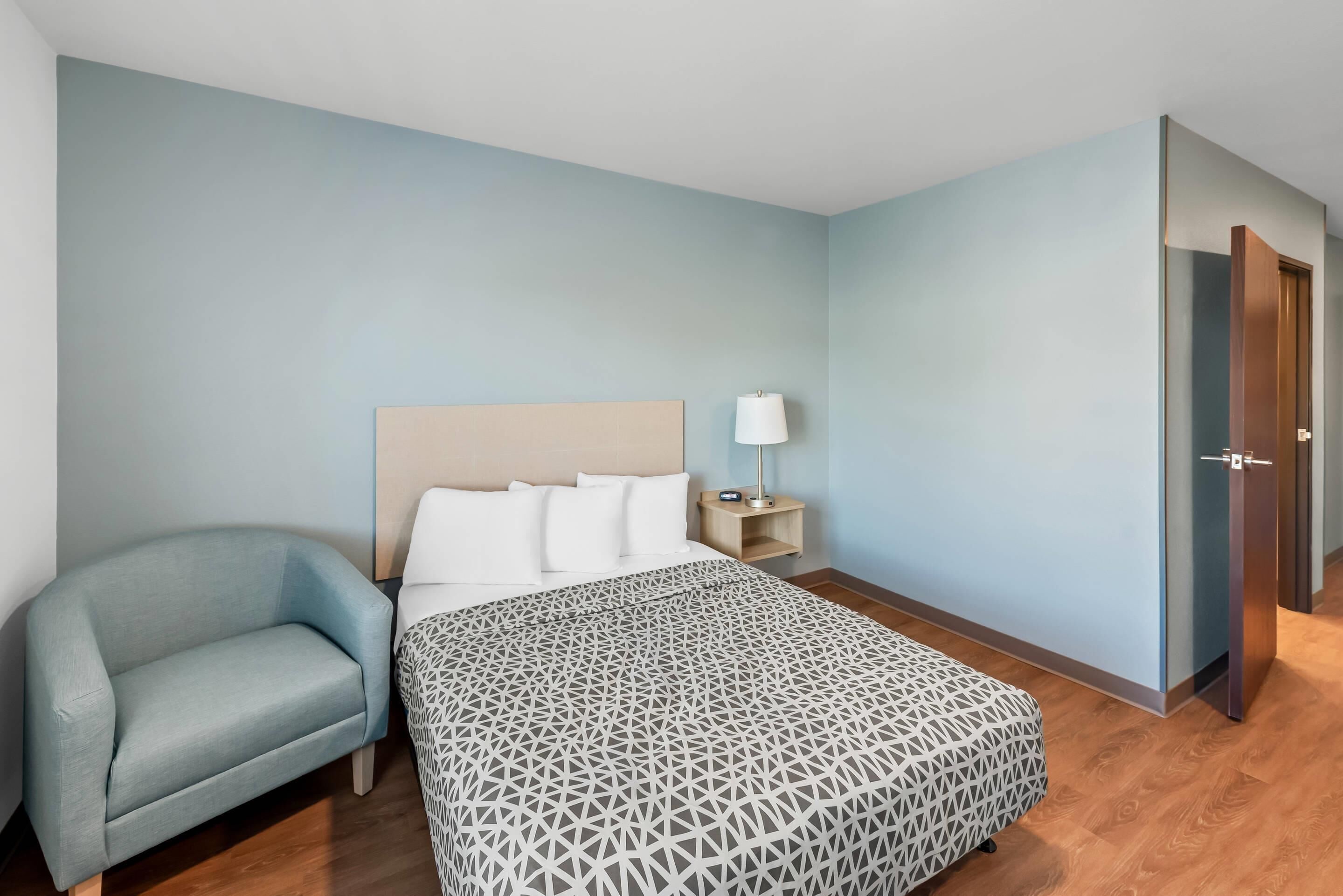 A hotel room with a bed, a bedside table with a lamp, a chair, and a door.