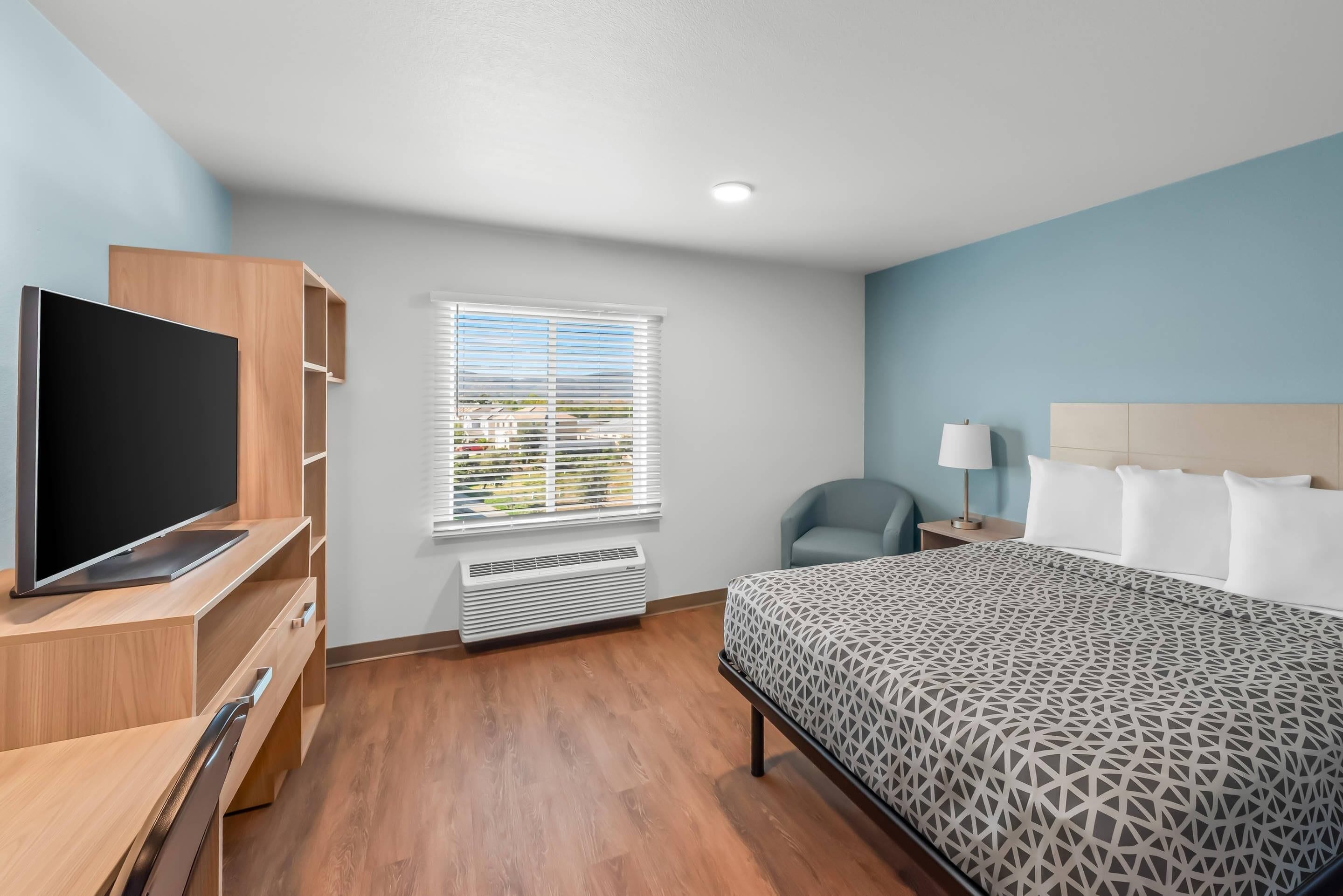 A hotel room with a bed, a TV, a desk, a chair, and a window with blinds.
