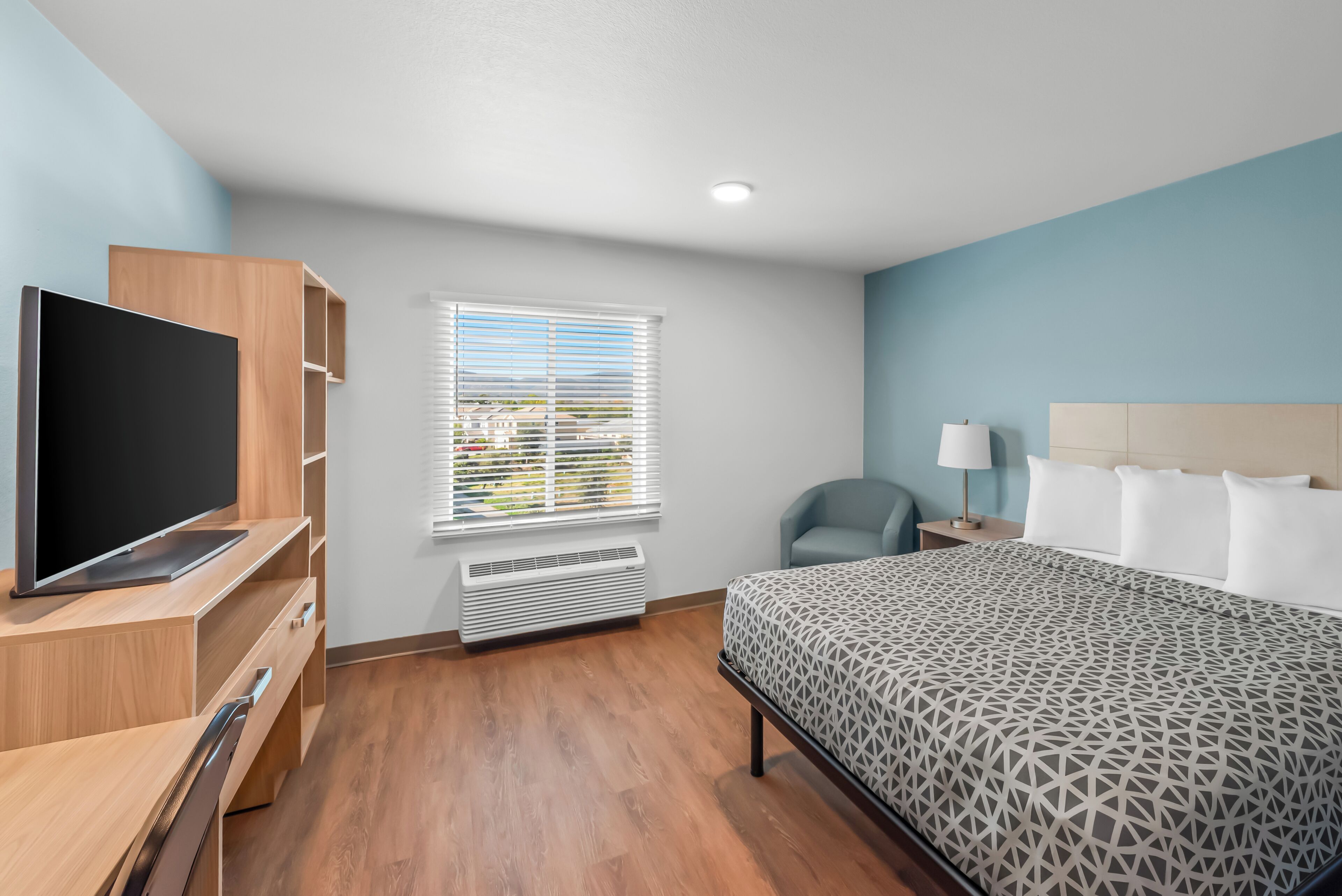 A hotel room with a bed, a TV, a desk, a chair, a window with blinds, and a nightstand with a lamp.