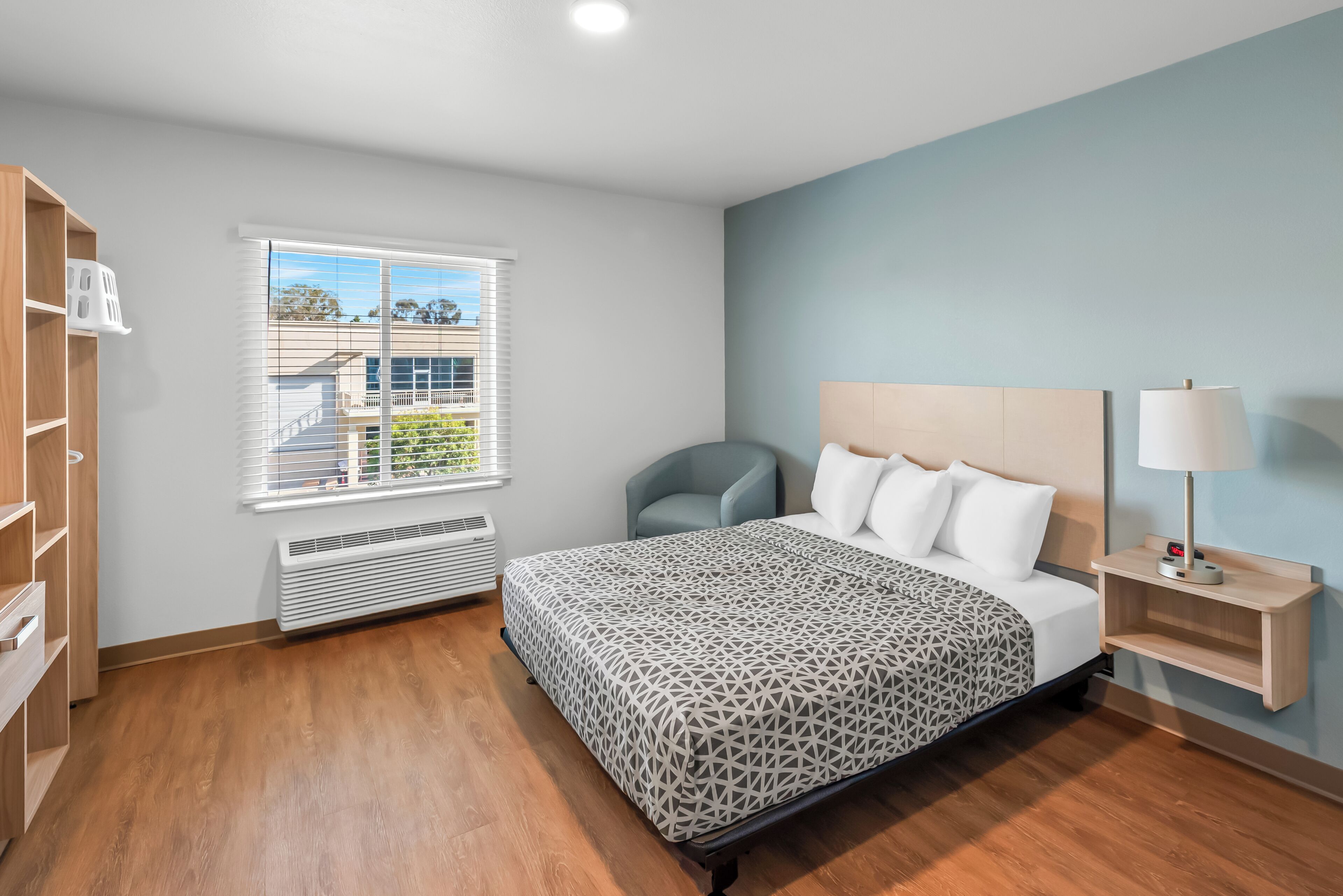 Quarto de hotel com cama, cadeira, mesa de cabeceira, luminária, aquecedor e janela com persianas.