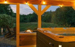 A hot tub under a wooden pergola with string lights, overlooking a grassy area with trees.