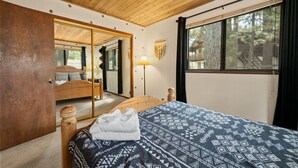 A bedroom with a wooden ceiling, a large mirror, a bed with a patterned blanket, a bedside lamp, and a window with a view of trees.