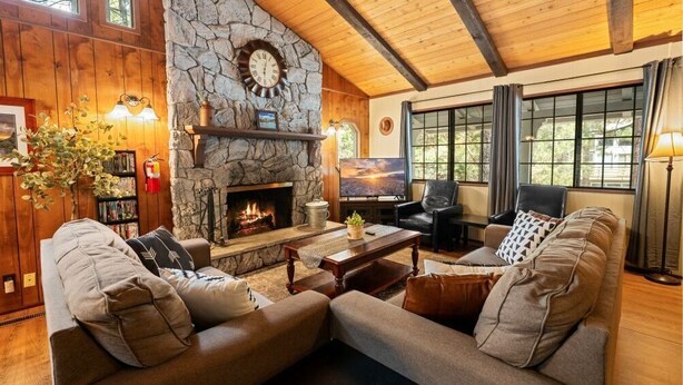 A cozy living room with a stone fireplace, wooden ceiling beams, and large windows.