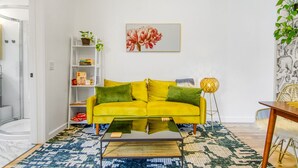 Un canapé jaune vif agrémenté de coussins verts, un tapis à motifs, une table basse en verre, une bibliothèque remplie de livres et de plantes, et une œuvre d’art florale encadrée accrochée au mur.