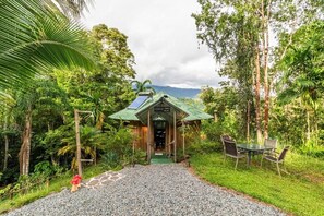 A rustic wooden building with a green roof, surrounded by lush greenery and a gravel path leading to a table and chairs.