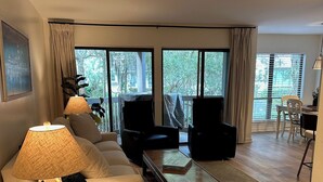 Un salon moderne comprenant un canapé, deux fauteuils, une table basse et une lampe. De grandes fenêtres offrent une vue sur les arbres et un balcon.