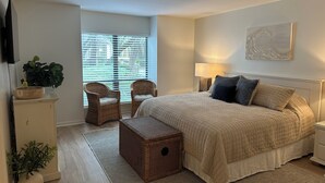 Une chambre avec un grand lit, deux fauteuils en osier, une table de chevet avec une lampe, une petite commode en bois et une plante en pot.