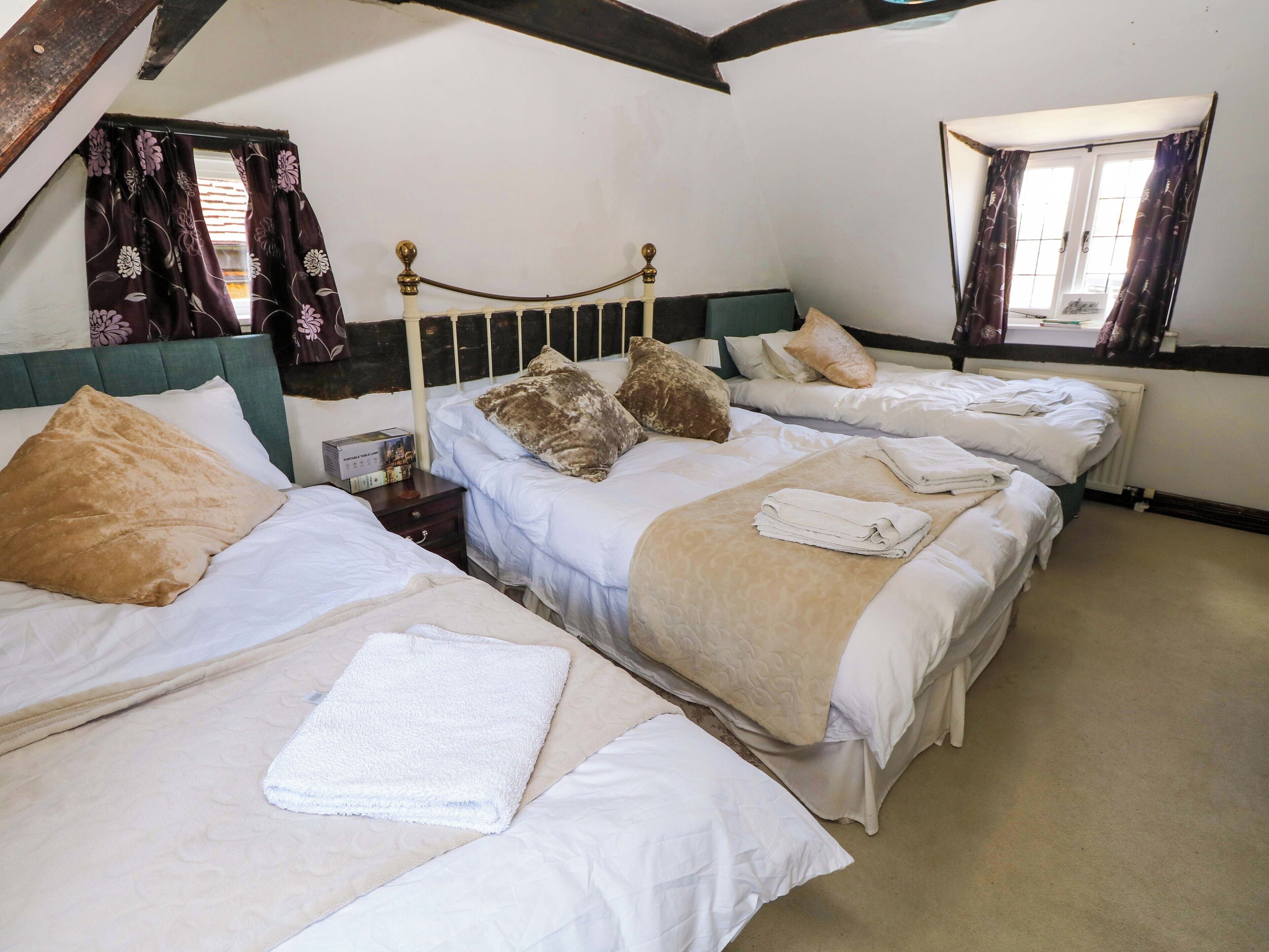 A hotel room with two single beds, each with a pillow and a folded towel. There is a nightstand with a lamp and a box of tissues. The room has a wooden ceiling and a window with curtains.