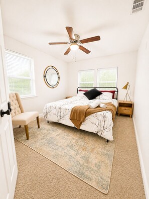 A bedroom with a bed, a chair, a ceiling fan, and two windows with blinds.