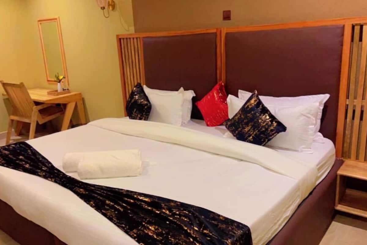 A neatly made bed with a white comforter, a black and gold throw blanket, and several pillows in various colors. There is a wooden headboard, a small wooden table with a chair, and a mirror on the wall.
