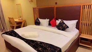 Una cama bien hecha con una colcha blanca, un plaid negro y dorado, y varios almohadones de diferentes colores. Se observa un cabecero de madera, una mesita de madera con una silla, y un espejo en la pared.