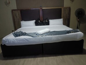 A neatly made bed with white linens, a dark headboard, and a black fan to the right.