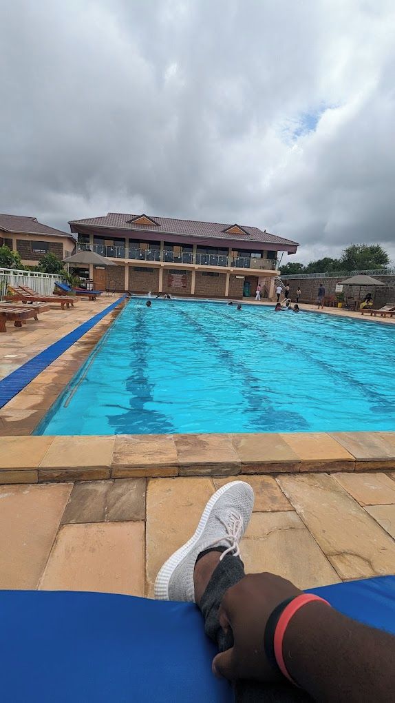Una piscina con una persona recostada en una tumbona, un edificio al fondo y un cielo nublado.
