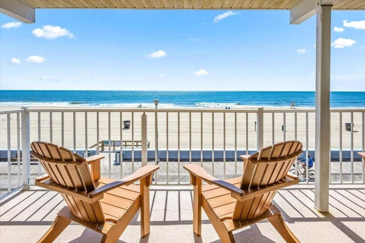A balcony with two wooden chairs and a table, overlooking a beach with a clear view of the ocean and a sandy shore.